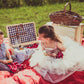 A picnic blanket in a field with a picnic basket and two kids sitting their holding soft pretzels and cloth napkins. Behind them is an apple crate with a basket some games. Created in the orchard of Nystrom Orchard in Worthington Minnesota.