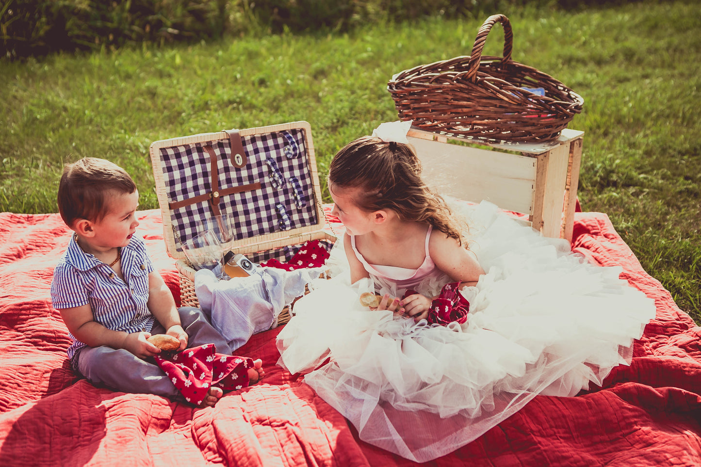 A picnic blanket in a field with a picnic basket and two kids sitting their holding soft pretzels and cloth napkins. Behind them is an apple crate with a basket some games. Created in the orchard of Nystrom Orchard in Worthington Minnesota.