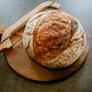 Rustic loaf of plain sourdough bread on a wooden cutting board with a wooden bow knife on a dark surface. Baked in the kitchen of Nystrom Orchard in Worthington Minnesota.