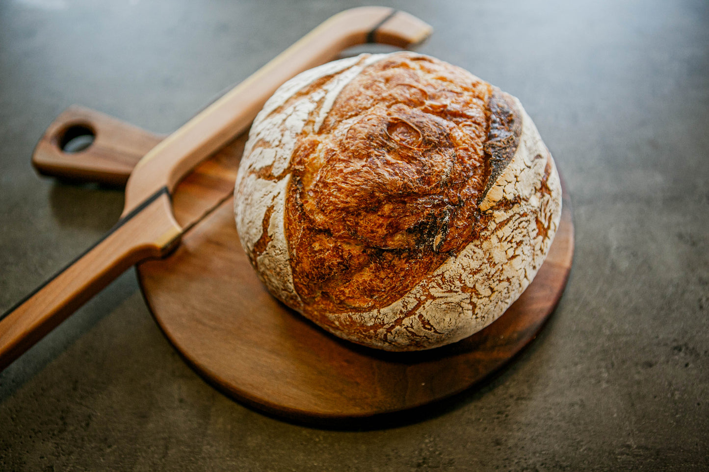 Rustic loaf of plain sourdough bread on a wooden cutting board with a wooden bow knife on a dark surface. Baked in the kitchen of Nystrom Orchard in Worthington Minnesota.