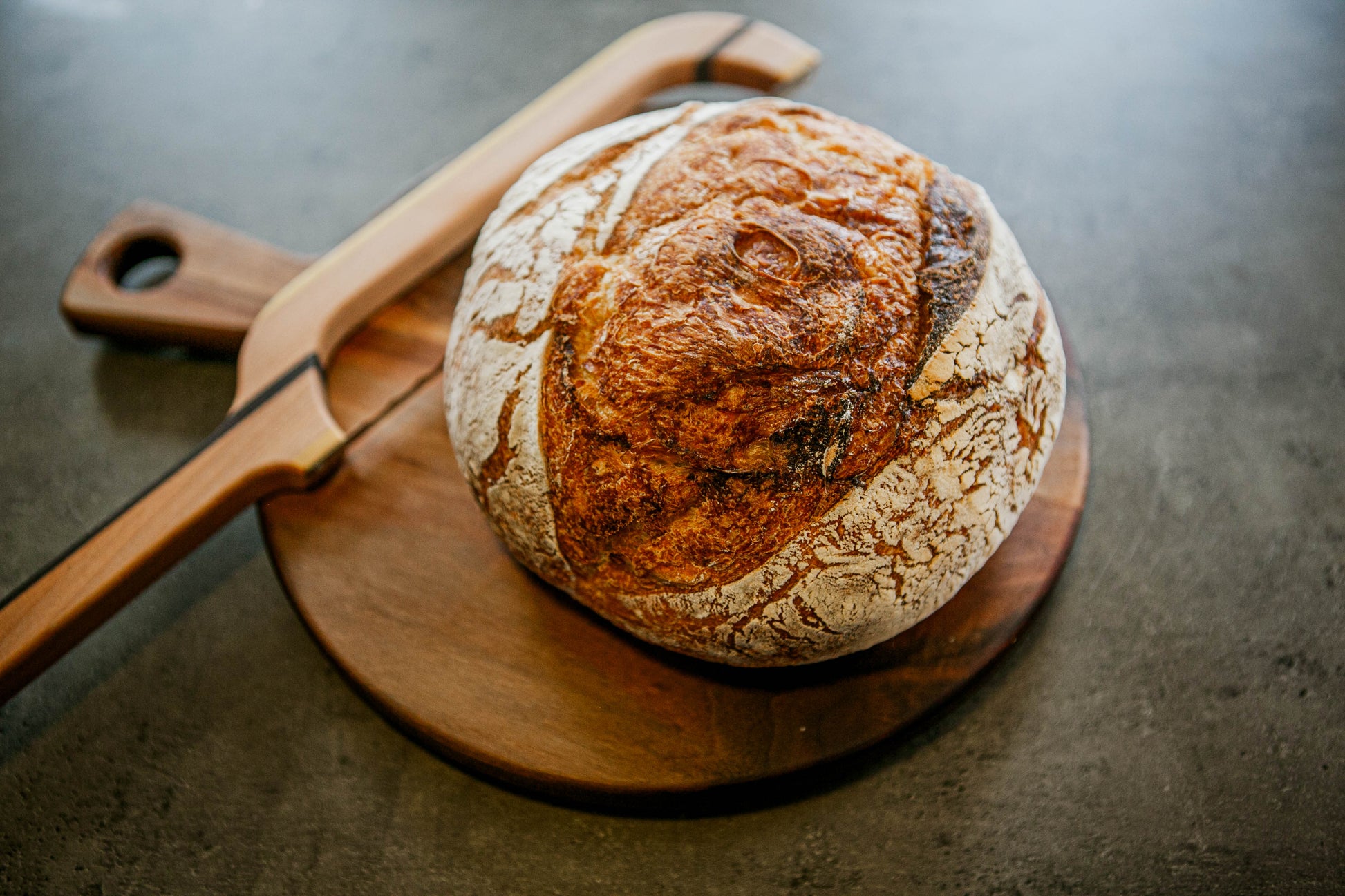 Rustic loaf of plain sourdough bread on a wooden cutting board with a wooden bow knife on a dark surface. Baked in the kitchen of Nystrom Orchard in Worthington Minnesota.