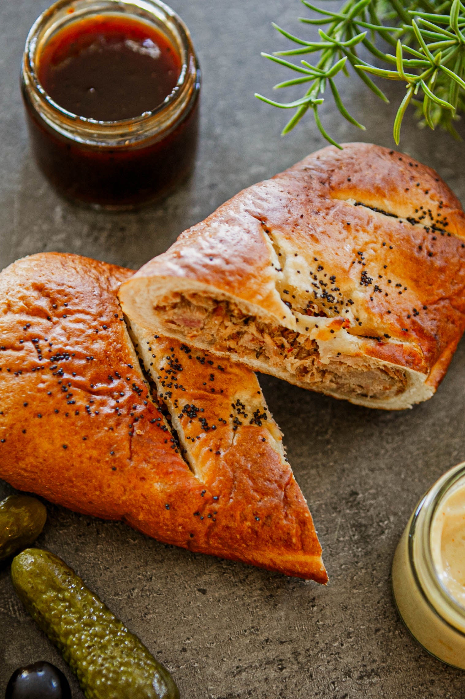 A photo showing a frozen artisan pretzel with smoked pulled pork filling, along with condiments and a pickle on a dark background. Made in the kitchen of Nystrom Orchard in Worthington Minnesota.