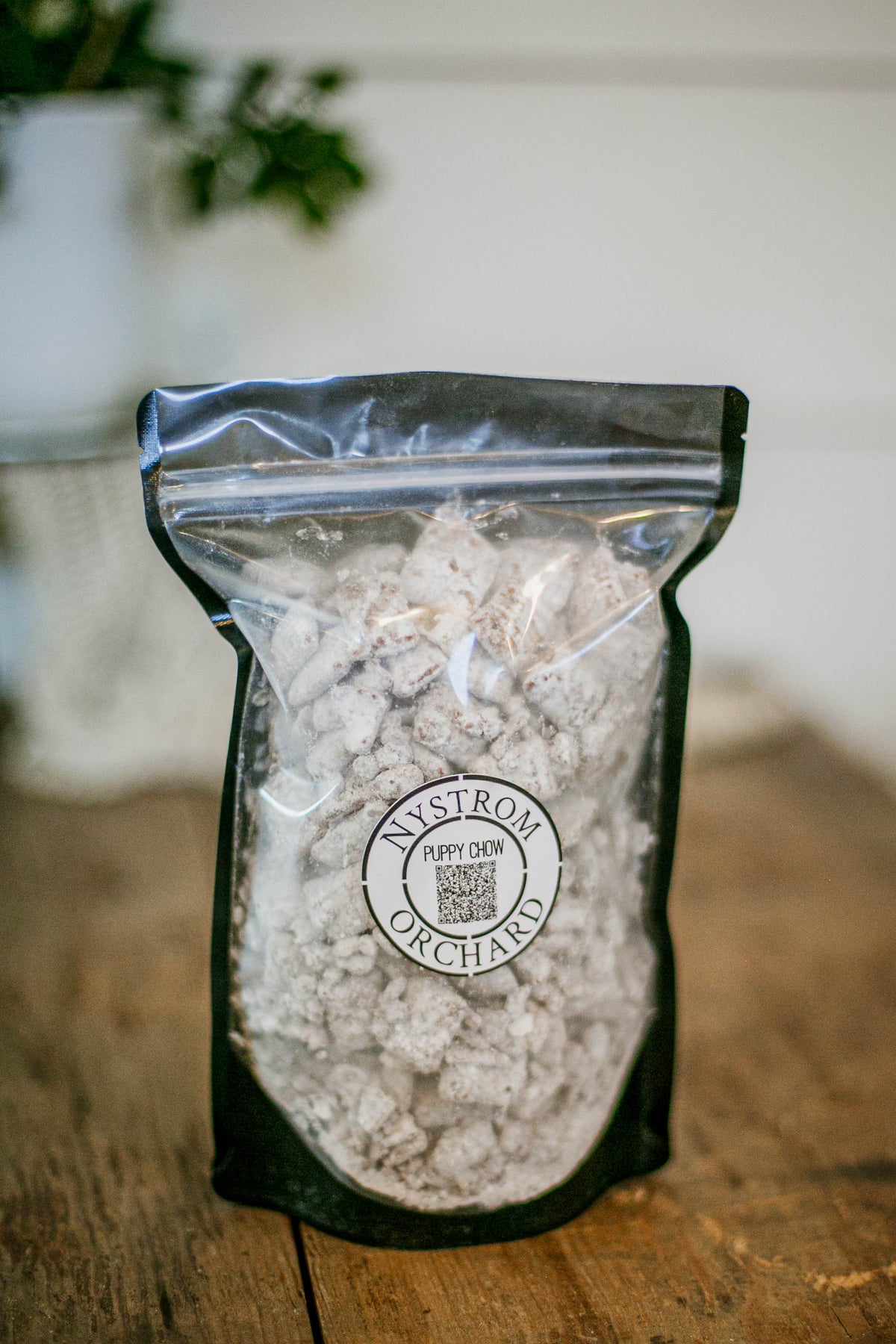 Bag of snack mix including rice chex, peanut butter, chocolate, and powdered sugar on a wooden table with a blurry background. Made in the kitchen of Nystrom Orchard in Worthington Minnesota.