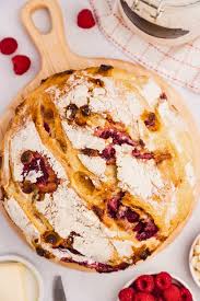 A whole rustic loaf of sourdough bread on a cutting board. There are raspberries and white chocolate beside the loaf. The loaf is baked at Nystrom Orchard in Worthington Minnesota. 