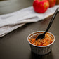 Single serving of scalloped apples in a metal dish with a black spoon with a dark surface and apples in the background. Baked in the kitchen of Nystrom Orchard in Worthington Minnesota.