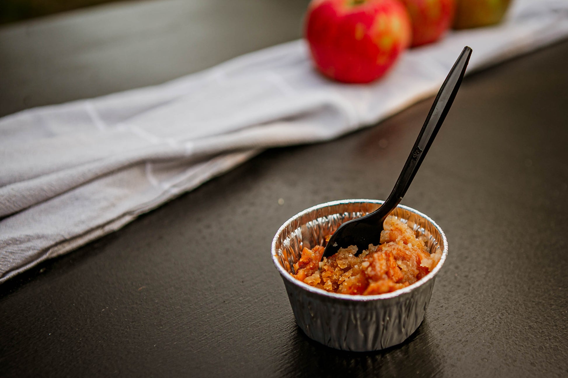Single serving of scalloped apples in a metal dish with a black spoon with a dark surface and apples in the background. Baked in the kitchen of Nystrom Orchard in Worthington Minnesota.