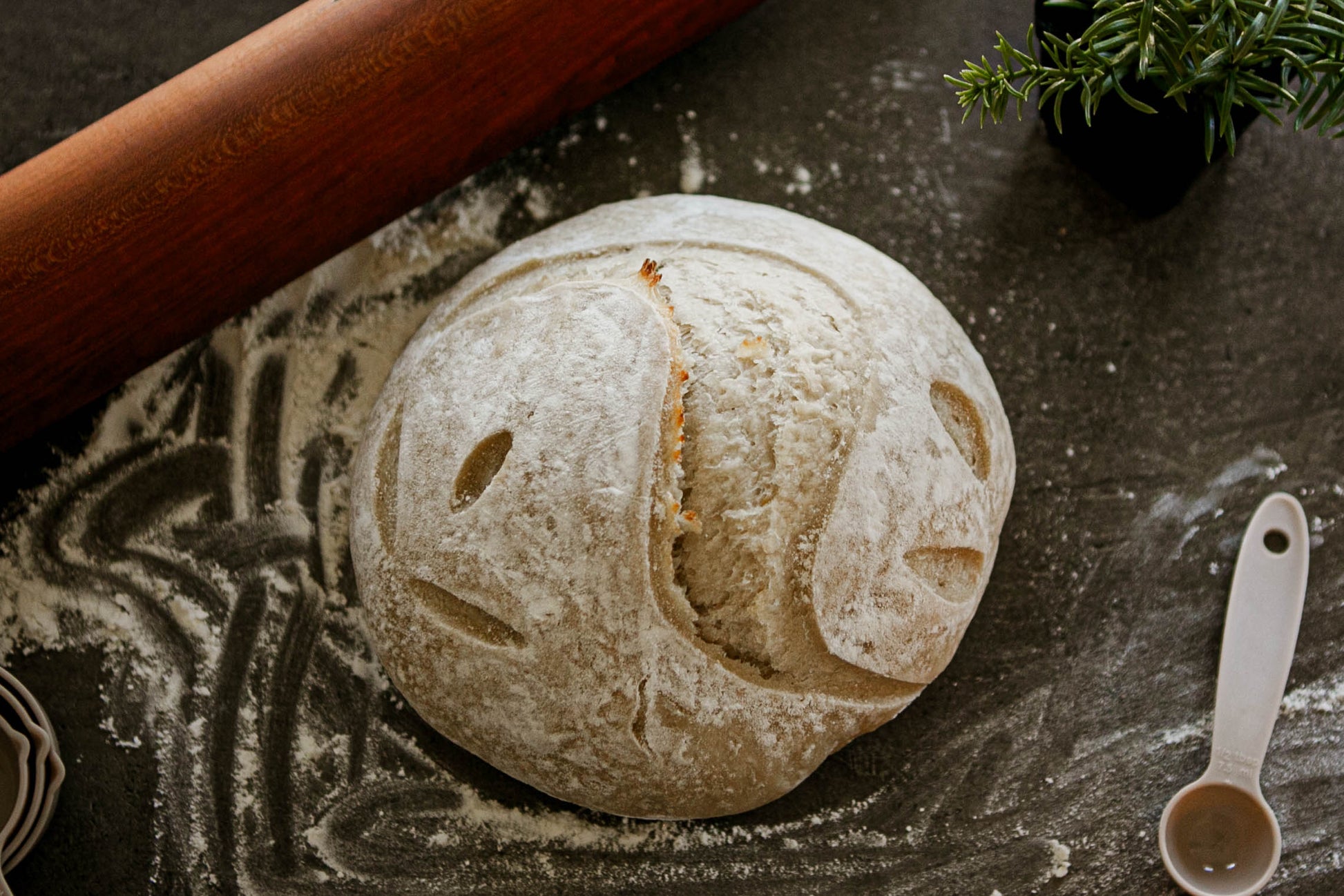 Rustic loaf of rosemary sea salt sourdough bread on a floured surface with a green plant, measuring spoon and rolling pin beside it. Baked in the kitchen at Nystrom Orchard in Worthington Minnesota.