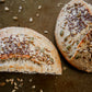 Two halves of a loaf of rustic hearty sourdough bread with seeds on a dark surface with seeds scattered around them. Baked in the kitchen at Nystrom Orchard in Worthington Minnesota.