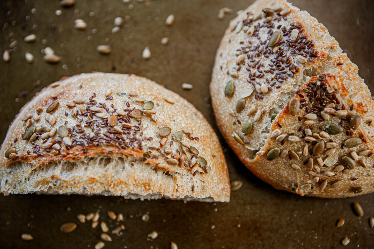 Two halves of a loaf of rustic hearty sourdough bread with seeds on a dark surface with seeds scattered around them. Baked in the kitchen at Nystrom Orchard in Worthington Minnesota.