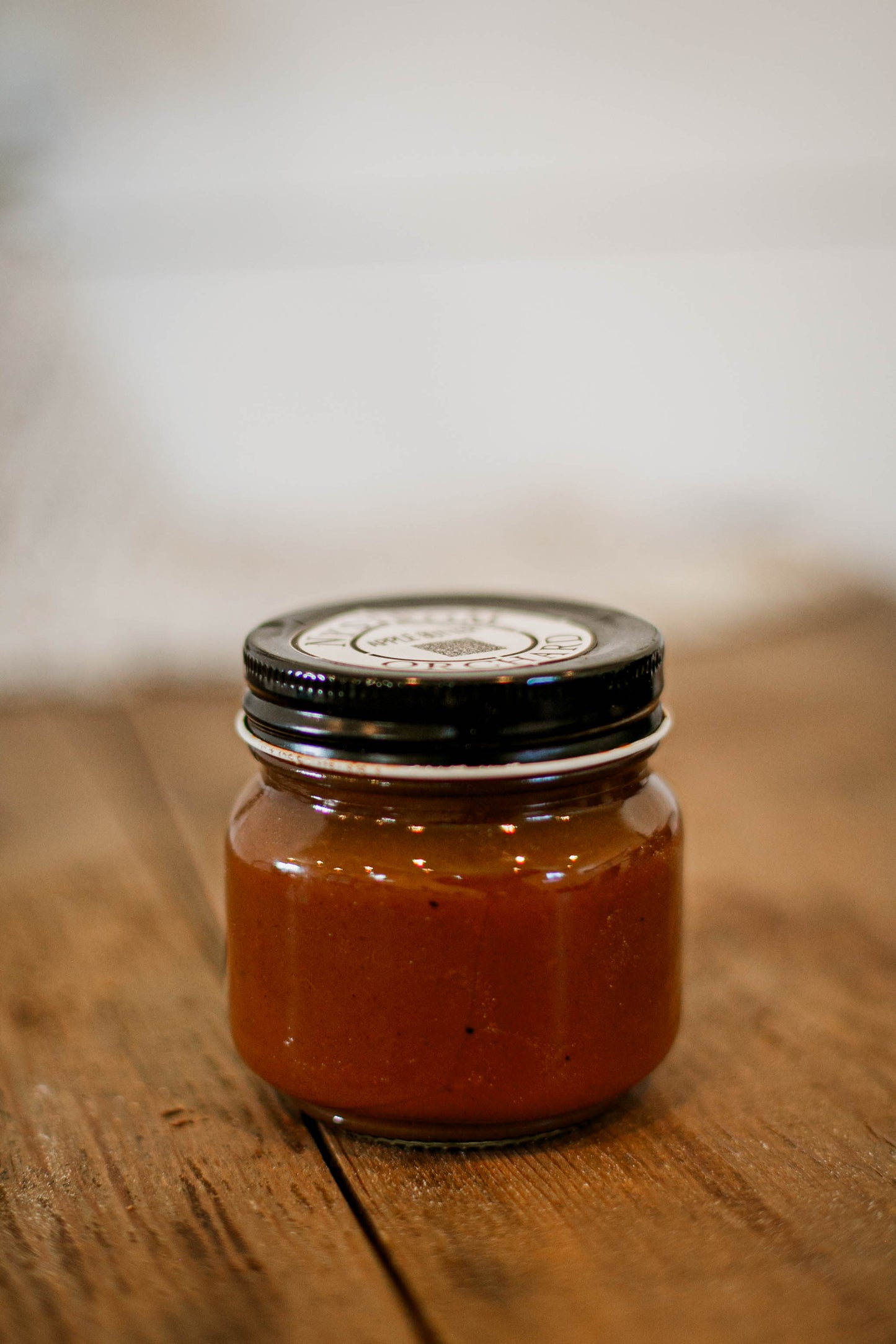 Glass 8 oz jar of apple butter with a black lid on a wooden surface. Made in the kitchen of Nystrom Orchard in Worthington Minnesota.