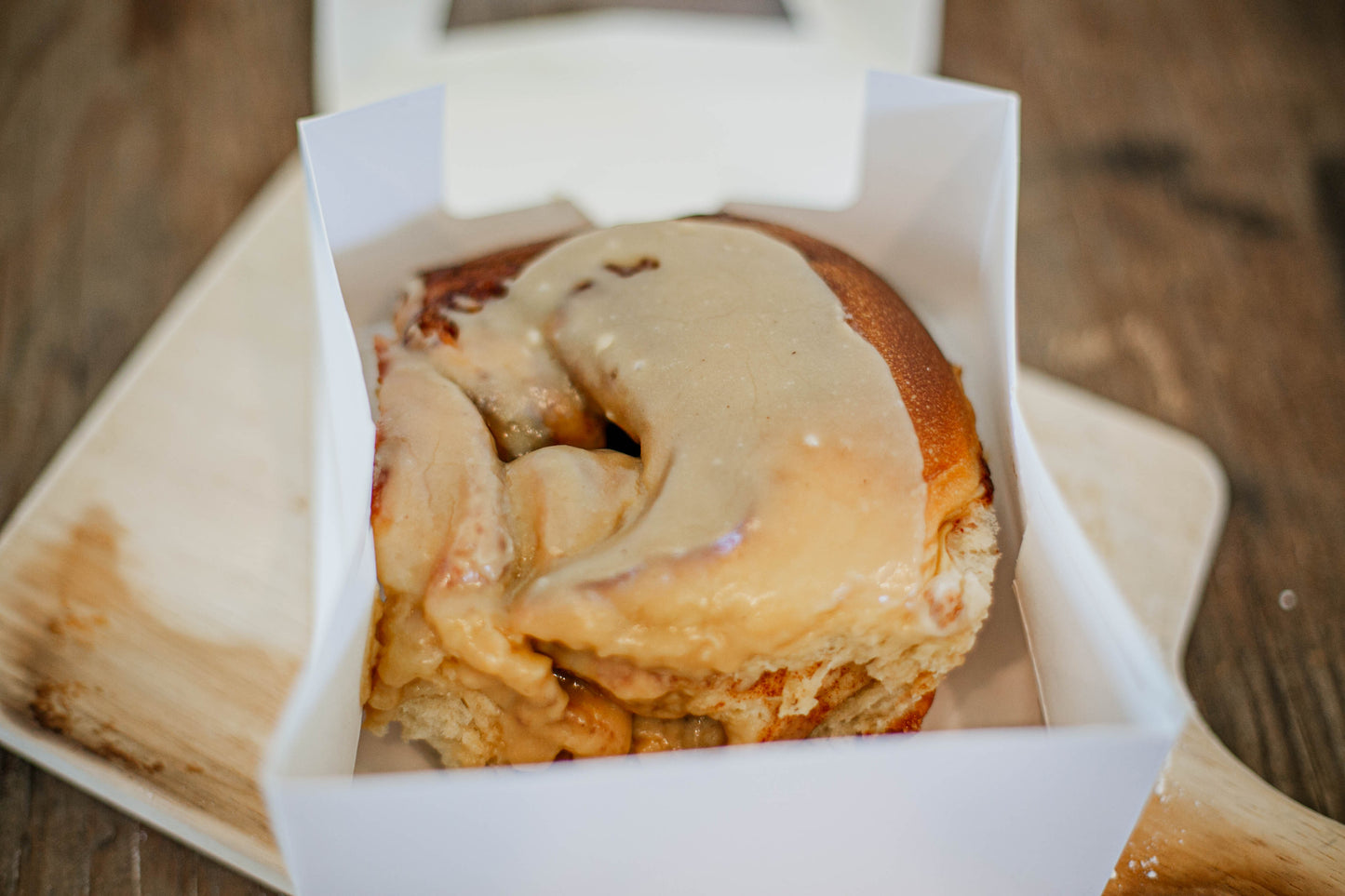 Single sourdough cinnamon roll in a white box on a cutting board with a wooden background. Baked in the kitchen of Nystrom Orchard in Worthington Minnesota.