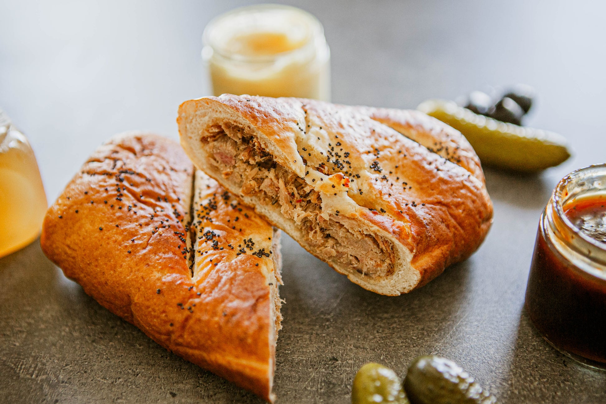 A soft pretzel wrap cut in half to show its full of smoked pulled pork surrounded by bbq, honey mustard and pickles on a dark surface. Baked in the kitchen of Nystrom Orchard in Worthington Minnesota.