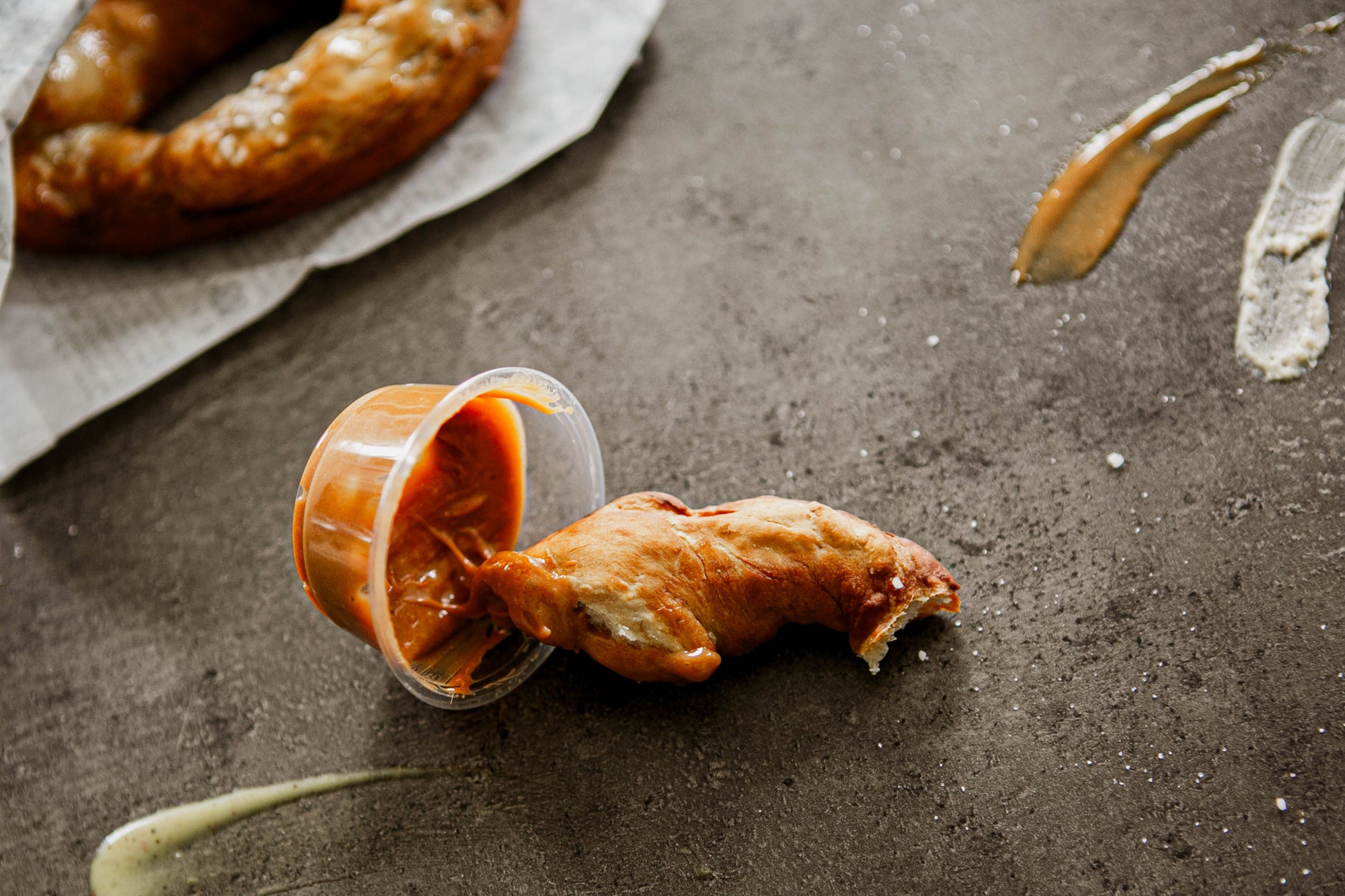 A piece of soft pretzel in a salted caramel dip container on a dark surface with another soft pretzel blurry in the background. Baked in the kitchen of Nystrom Orchard in Worthington Minnesota.