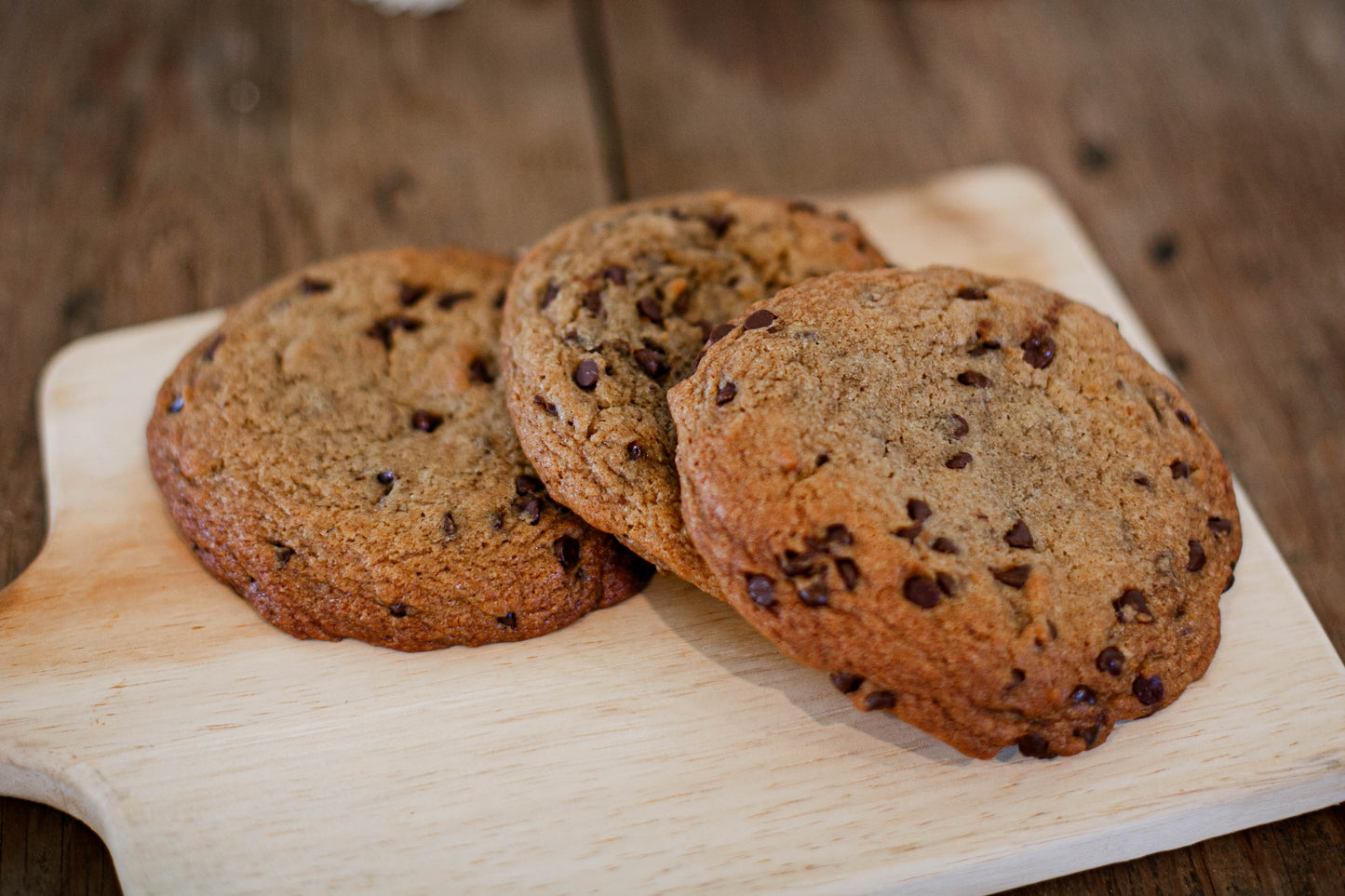Three chocolate chip cookies on a wooden cutting board with a blurred wooden background.  Baked in the kitchen of Nystrom Orchard in Worthington Minnesota.