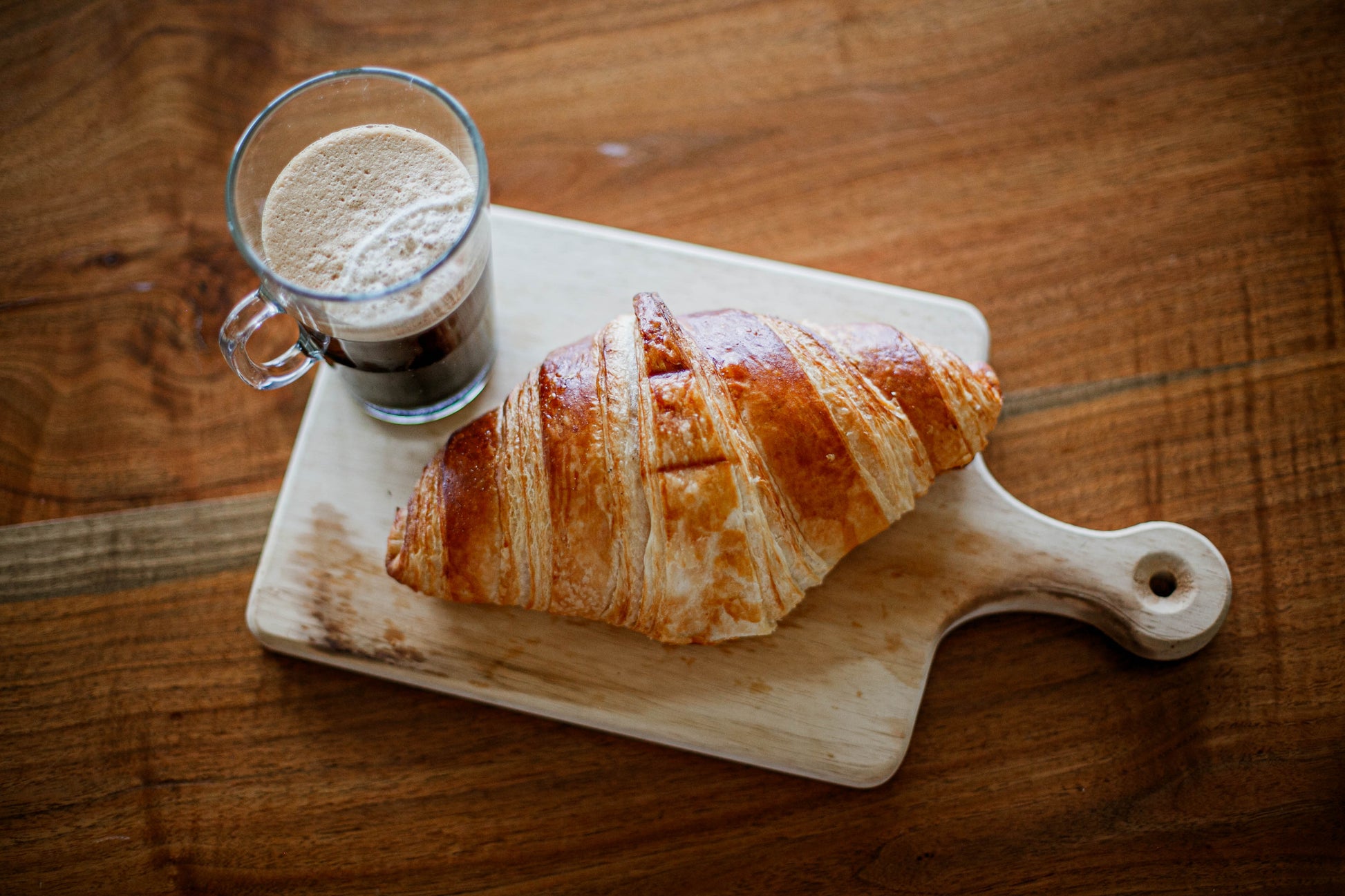 Homemade sourdough croissant on a wooden cutting board with a glass of coffee on a wooden surface. Baked in the kitchen of Nystrom Orchard in Worthington Minnesota.