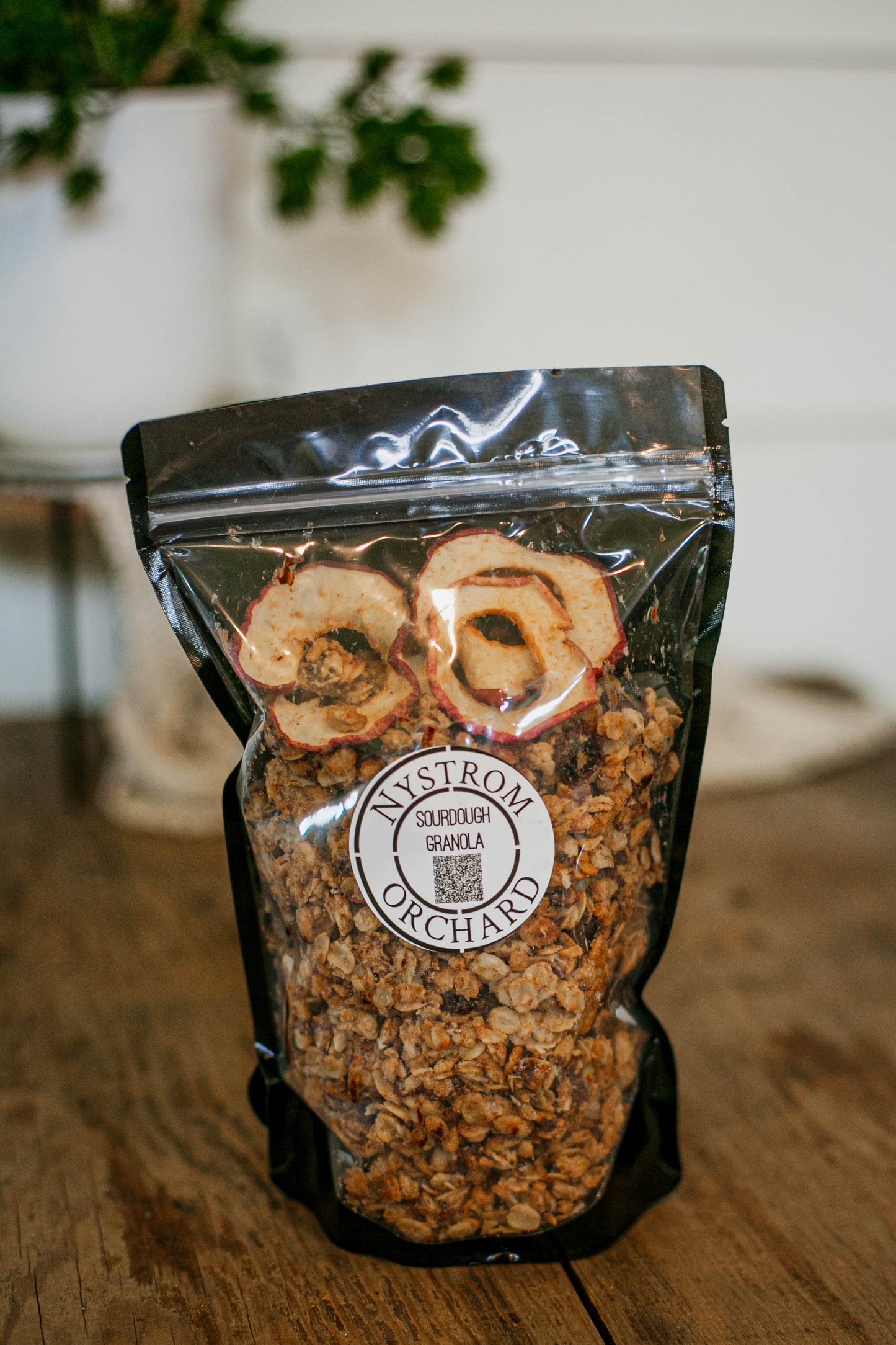 Bag of homemade sourdough granola with dried apples on a wooden surface, featuring the Nystrom Orchard brand. Made in the kitchen of Nystrom Orchard in Worthington Minnesota.