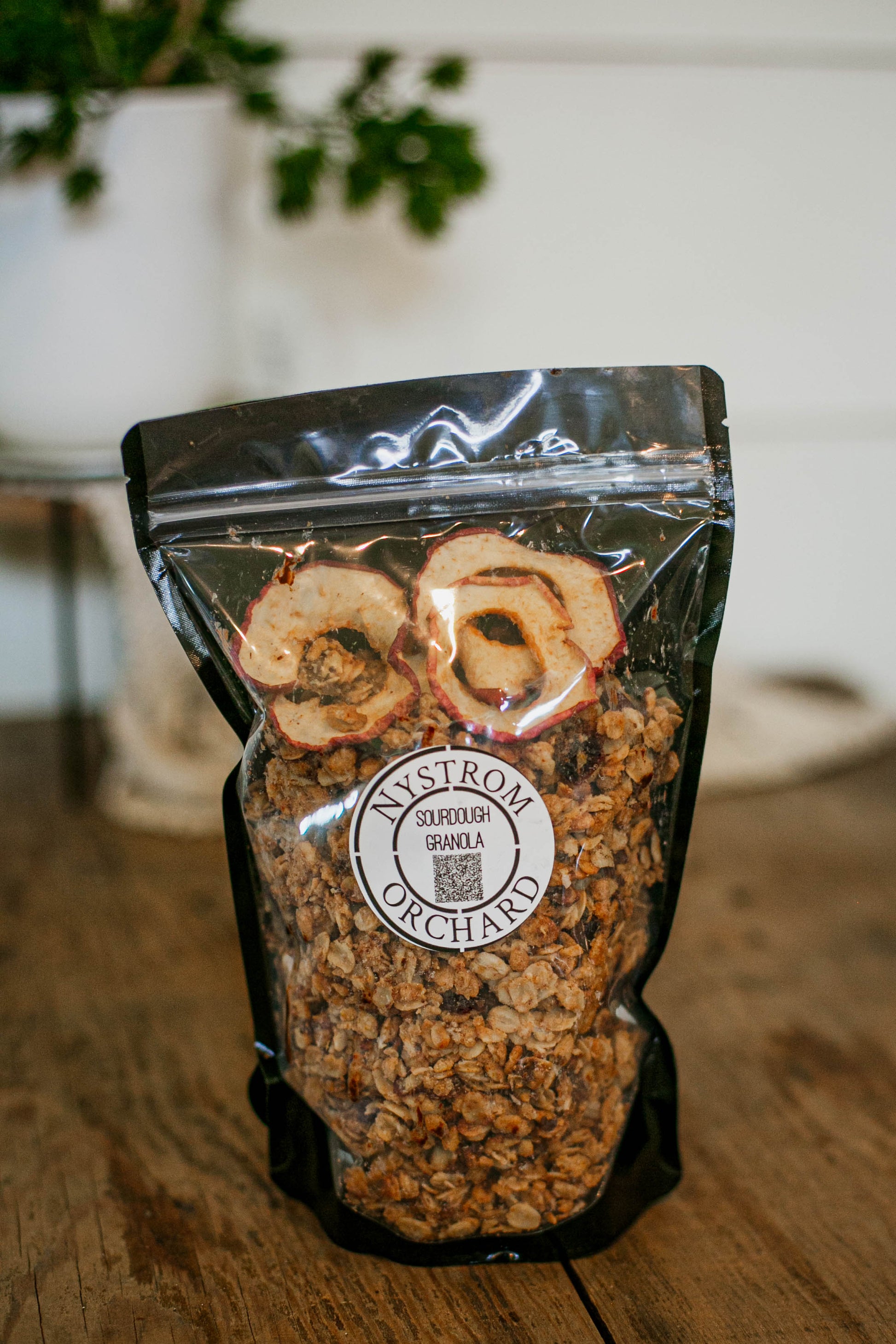Bag of homemade sourdough granola with dried apples on a wooden surface, featuring the Nystrom Orchard brand. Made in the kitchen of Nystrom Orchard in Worthington Minnesota.