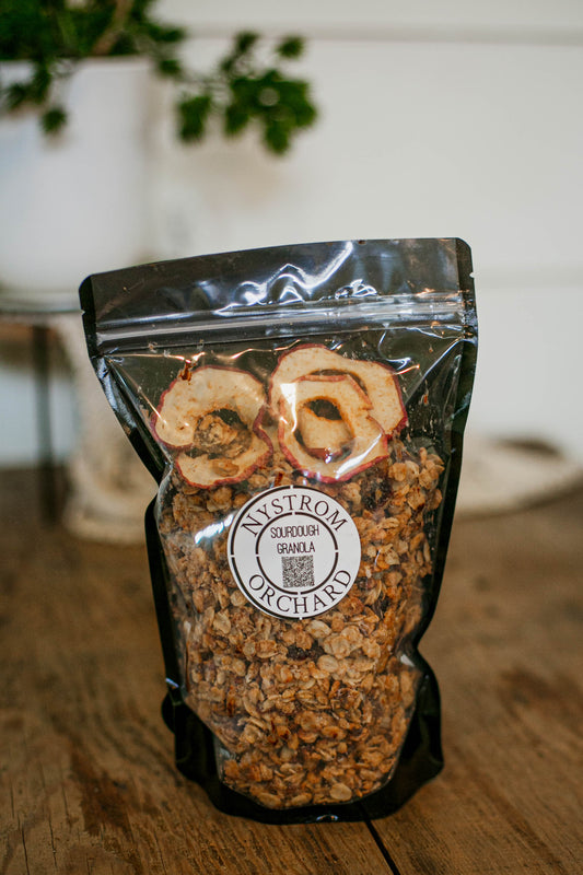 Bag of homemade sourdough granola with dried apples on a wooden surface, featuring the Nystrom Orchard brand. Made in the kitchen of Nystrom Orchard in Worthington Minnesota.