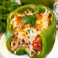A close-up of a stuffed green bell pepper on a white plate, filled with rice, ground meat, tomatoes, melted cheese, and parsley garnish, with another stuffed pepper blurred in the background. Made in the kitchen of Nystrom Orchard in Worthington Minnesota.