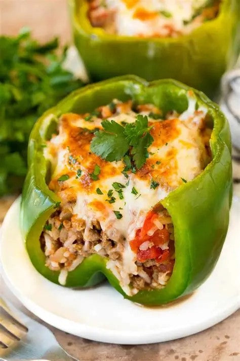 A close-up of a stuffed green bell pepper on a white plate, filled with rice, ground meat, tomatoes, melted cheese, and parsley garnish, with another stuffed pepper blurred in the background. Made in the kitchen of Nystrom Orchard in Worthington Minnesota.