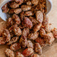 Toasted spiced almonds spilling out of a white cup on a wooden surface. Created by the kitchen at Nystrom Orchard in Worthington Minnesota. 