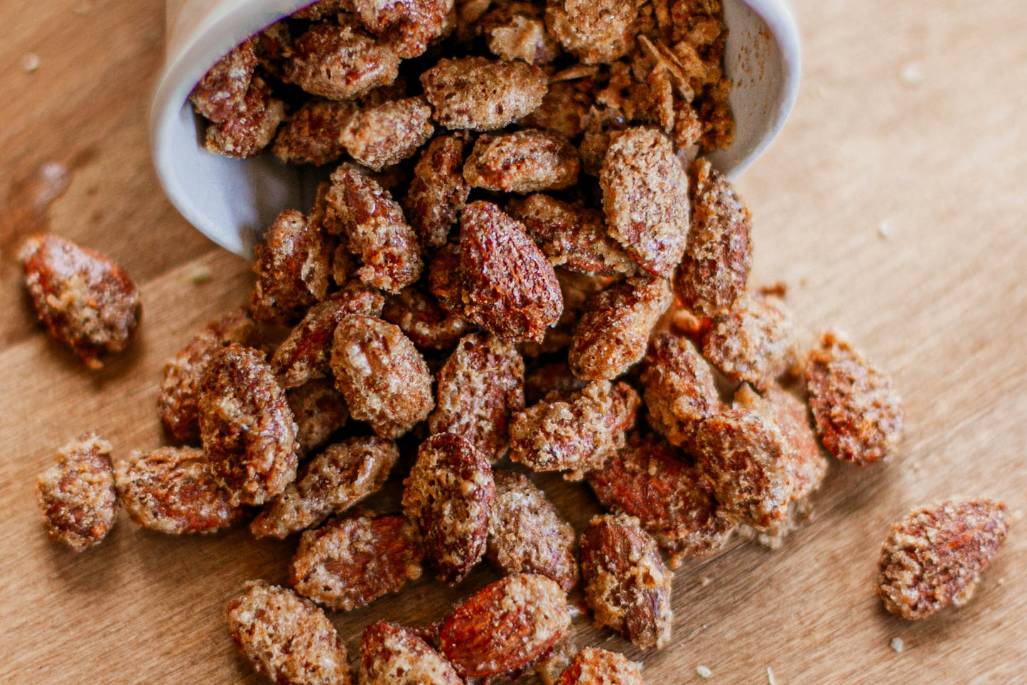 Toasted spiced almonds spilling out of a white cup on a wooden surface. Created by the kitchen at Nystrom Orchard in Worthington Minnesota. 