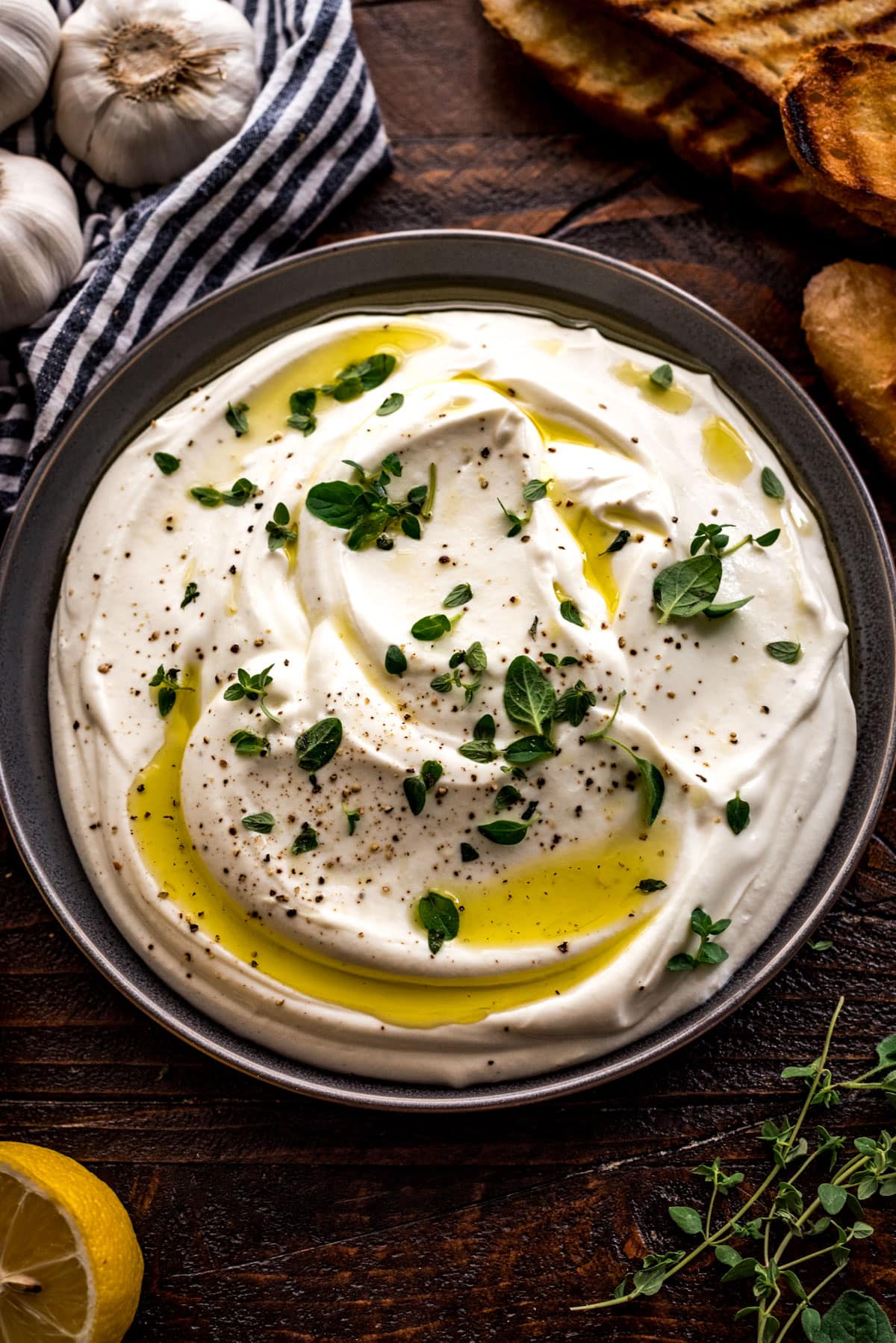 Creamy whipped ricotta with lemon and herbs on a dark bowl with herbs, garlic, lemons, and toasted bread lying beside on a wooden surface. Made in the kitchen of Nystrom Orchard in Worthington Minnesota.