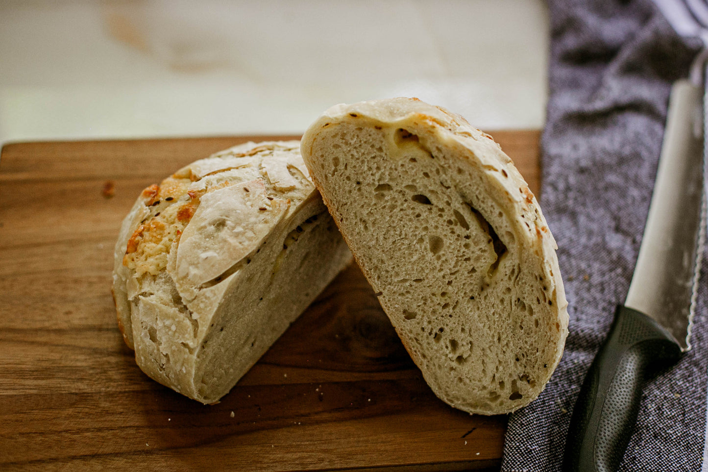 Sliced loaf of rustic white cheddar dill sourdough bread on a wooden cutting board with a knife and cloth in the background. Made in the kitchen of Nystrom Orchard in Worthington Minnesota. 