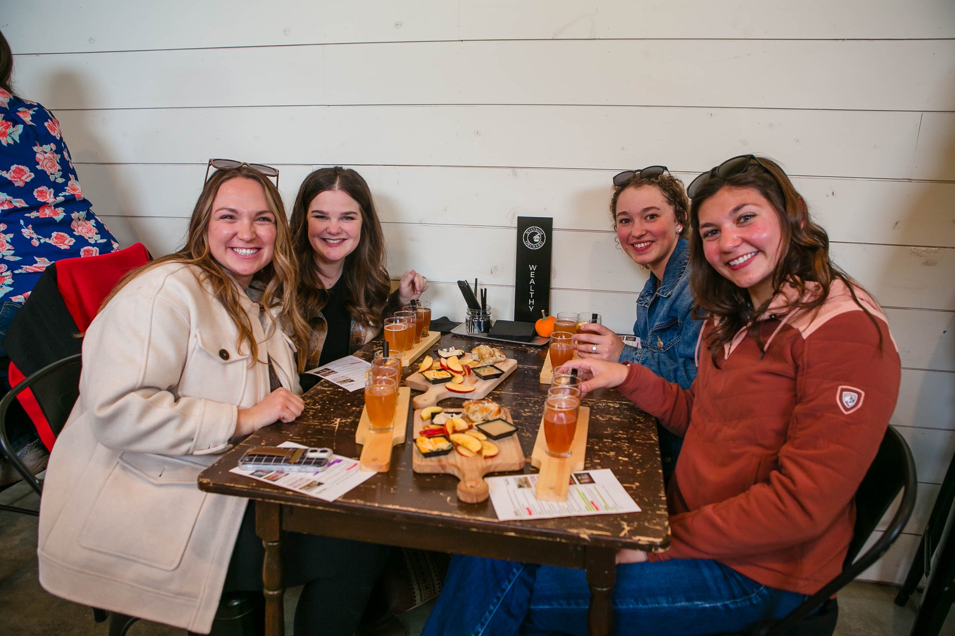 Four women sitting around a table with cider flights and food, smiling at the camera. Event hosted by Nystrom Orchard in Worthington Minnesota.
