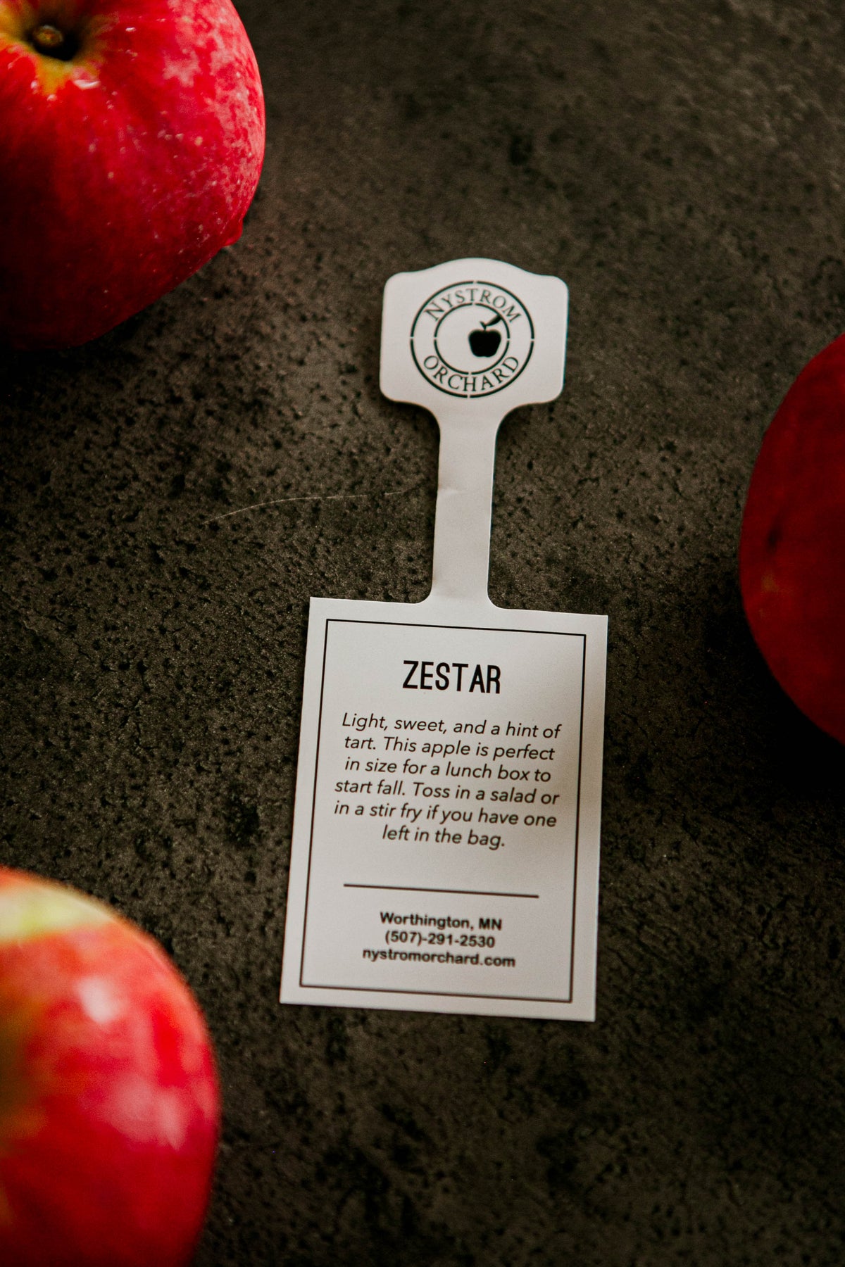 Zestar tag displayed on a textured surface with apples beside. Picked in Nystrom Orchard in Worthington Minnesota.