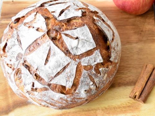 Rustic loaf of sourdough bread with a decorative pattern on a wooden surface with some cinnamon sticks and an apple beside it. Baked in the kitchen at Nystrom Orchard in Worthington Minnesota.