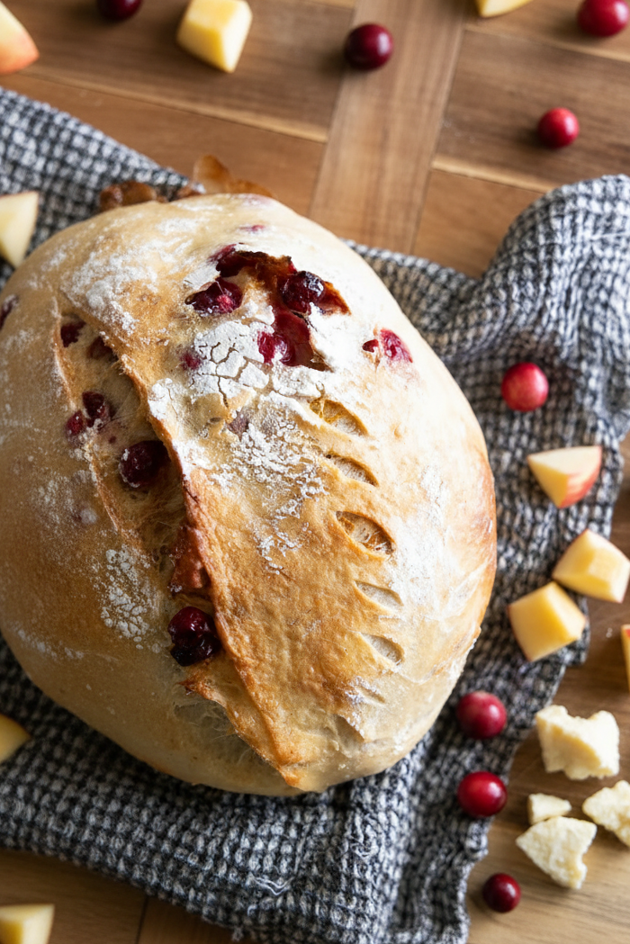 A rustic round artisan bread loaf with scored crust and cranberry garnish, resting on a gray checkered cloth on a wooden surface with apple slices, white cheddar, and whole cranberries scattered around. Baked in the kitchen Nystrom Orchard in Worthington Minnesota.