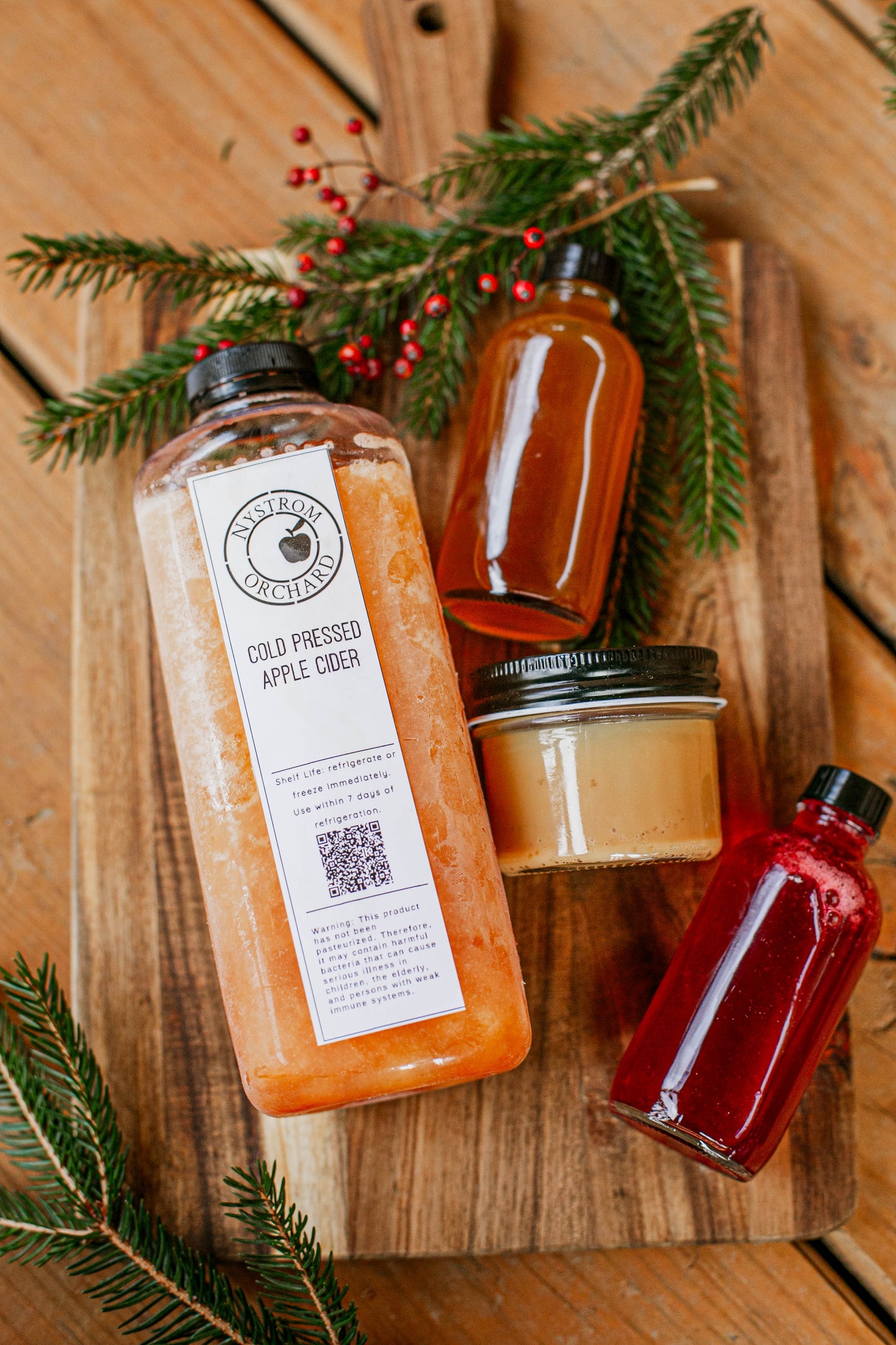 Bottle of cold pressed apple cider and cider syrup with salted caramel on a wooden board with greens and red berries. Made in the kitchen at Nystrom Orchard in Worthington Minnesota. 