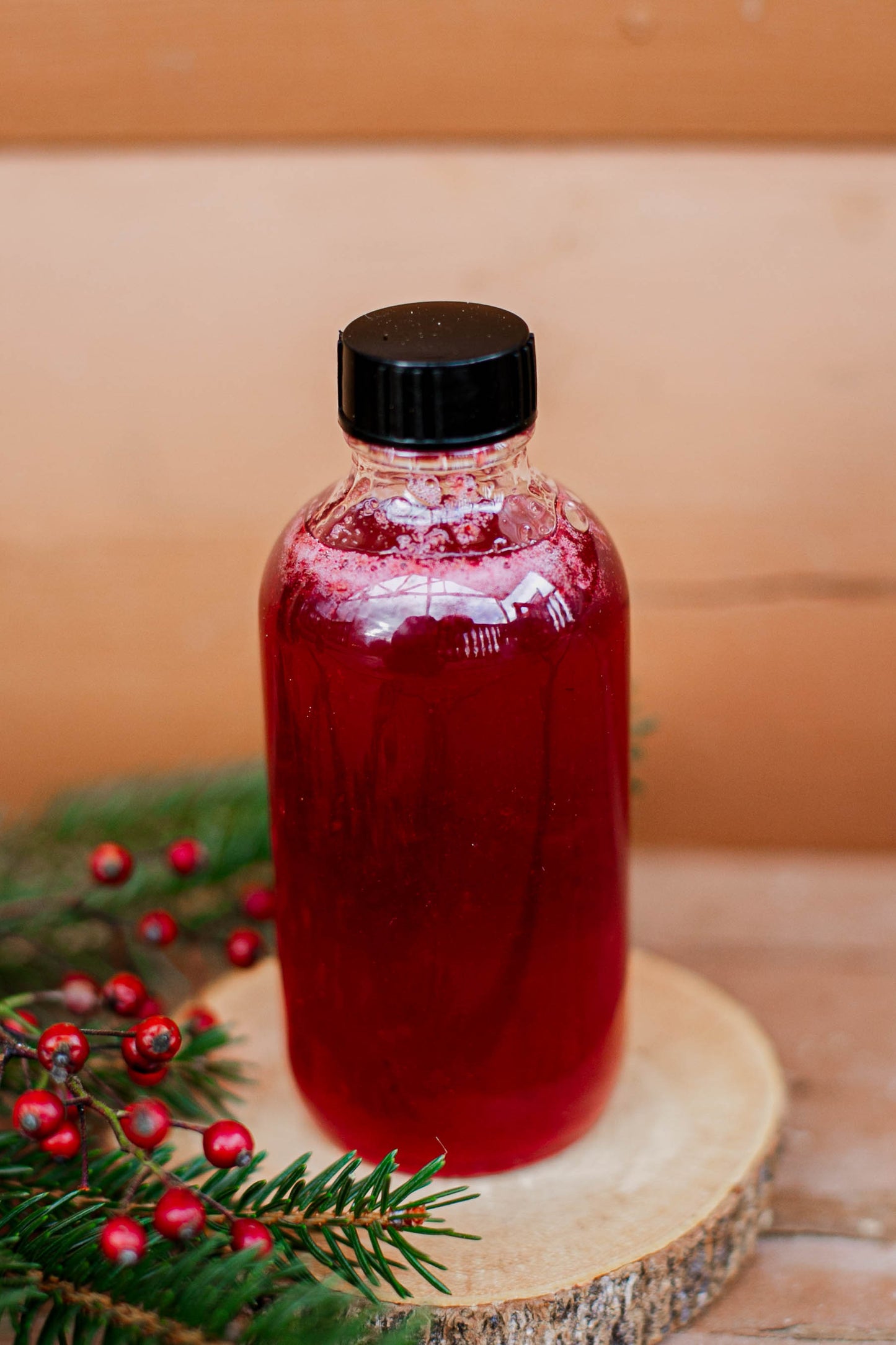 Bottle of cranberry vanilla syrup on a wooden surface with greenery and red berries. Made in the kitchen at Nystrom Orchard in Worthington Minnesota.