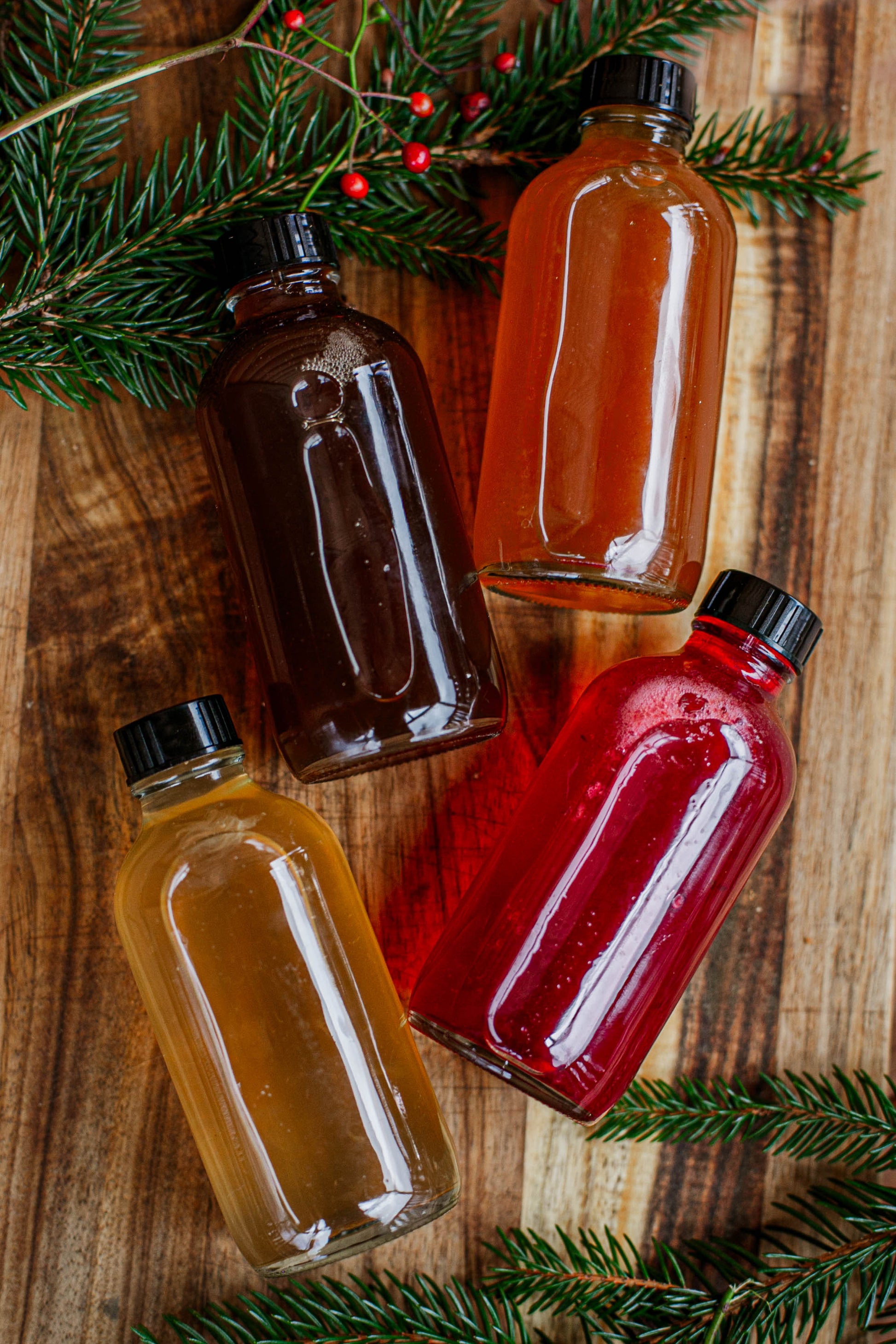 Four glass bottles of cider syrup on a wooden surface with greenery and red berries. Made in the kitchen at Nystrom Orchard in Worthington Minnesota.