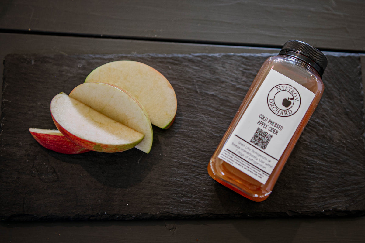 Photo of apple slices and a single serve bottle of cold pressed cider on a slate on a dark background. Pressed in the kitchen of Nystrom Orchard in Worthington Minnesota.