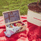 A picnic basket open on a red blanket in a field, filled with cold pressed cider and soft pretzels and accompanied by a wine glass and a basket with a blue and white checkered pattern on the inside. Beside there is another basket on an apple crate full of games. Packed in the kitchen of Nystrom Orchard in Worthington Minnestota.