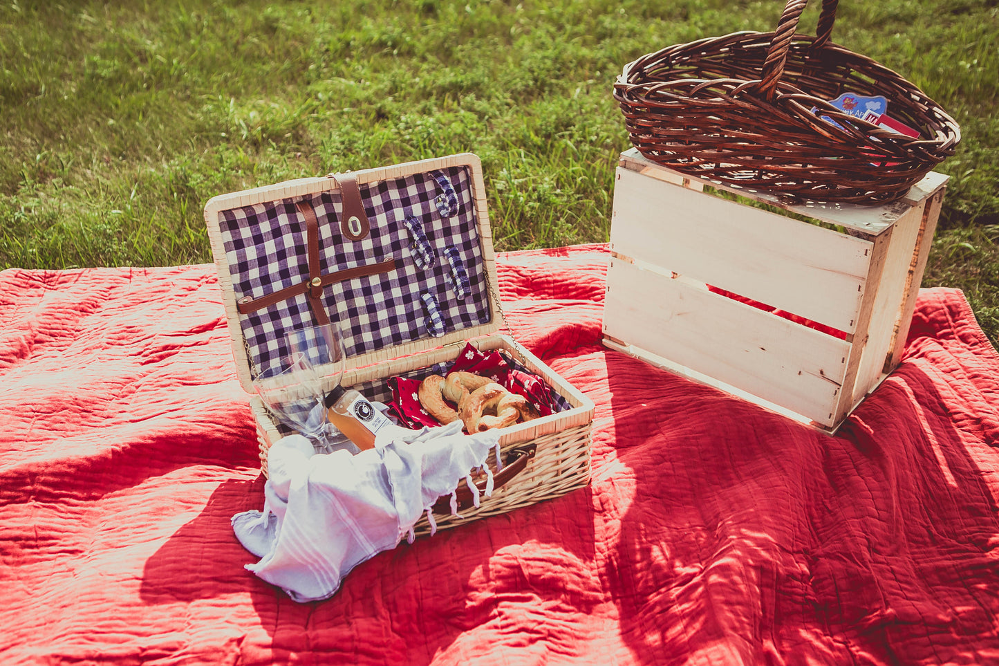 A picnic basket open on a red blanket in a field, filled with cold pressed cider and soft pretzels and accompanied by a wine glass and a basket with a blue and white checkered pattern on the inside. Beside there is another basket on an apple crate full of games. Packed in the kitchen of Nystrom Orchard in Worthington Minnestota.