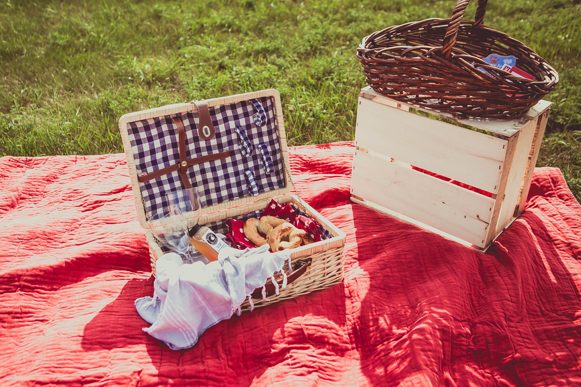 A picnic basket open on a red blanket in a field, filled with cold pressed cider and soft pretzels and accompanied by a wine glass and a basket with a blue and white checkered pattern on the inside. Beside there is another basket on an apple crate full of games. Packed in the kitchen of Nystrom Orchard in Worthington Minnestota.