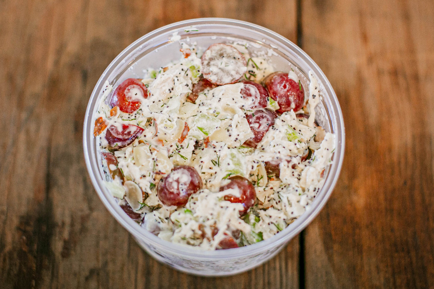 Container of chicken salad with grapes chicken celery, dill, poppy seeds on a wooden surface. Made in the kitchen of Nystrom Orchard in Worthington Minnesota.
