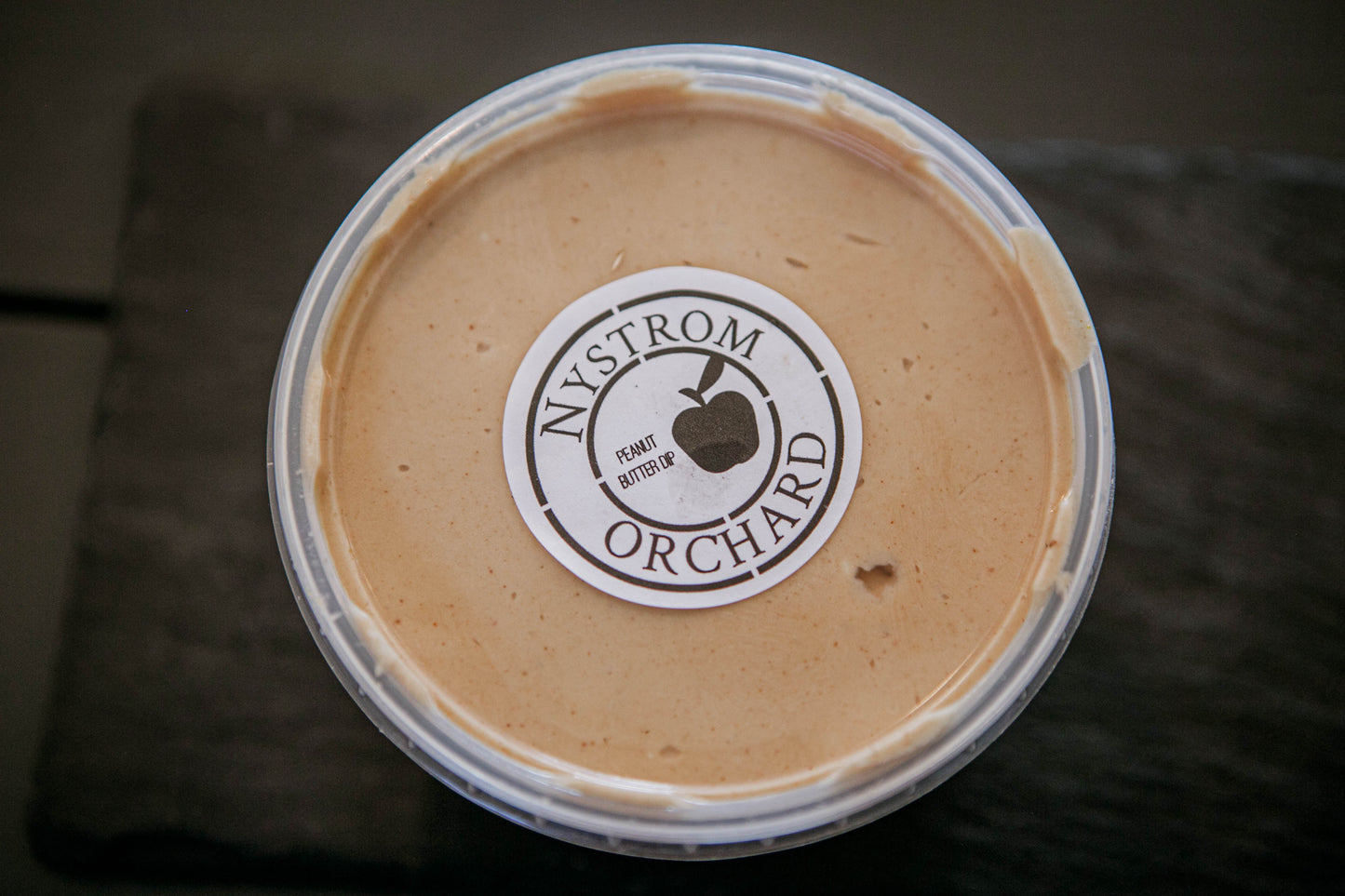 A plastic container of peanut butter cream cheese apple dip with a label that reads 'Nystrom Orchard' and has an image of an apple. Made in the kitchen of Nystrom Orchard in Worthington Minnesota.
