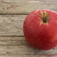 A single red apple with a slight sheen, placed on a wooden surface with visible grain and some paint residue. Picked in Nystrom Orchard in Worthington Minnesota.