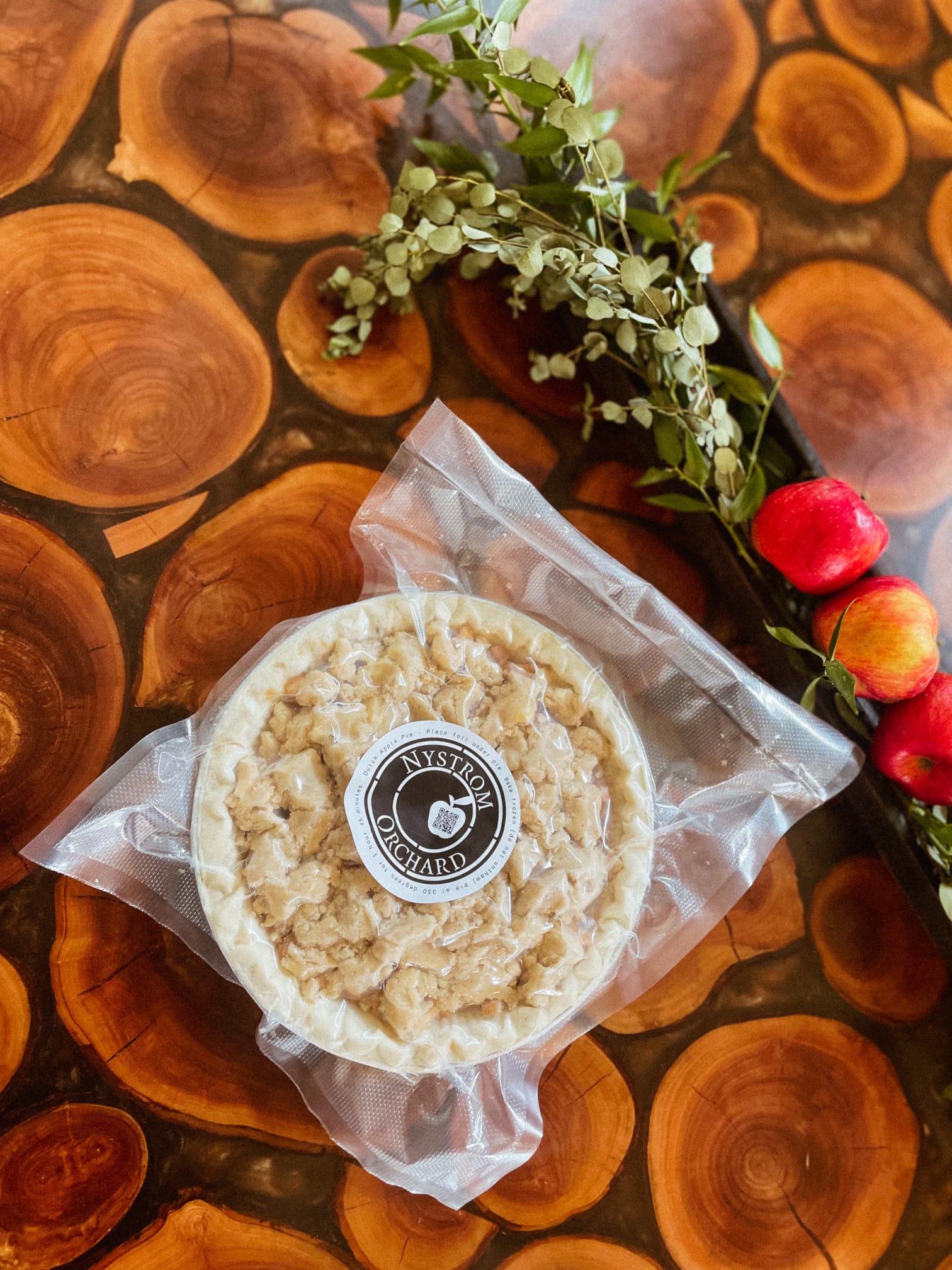 A packaged frozen apple pie with the Nystrom Orchard brand label on it on a wooden surface with decoration beside. Made in the kitchen of Nystrom Orchard in Worthington Minnesota.